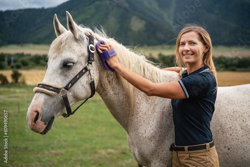 Young woman cleaning white gray horse with brush, her head next to animal