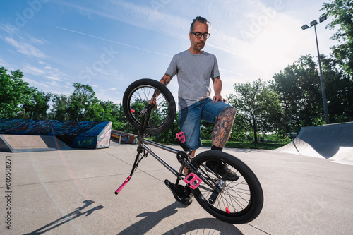 An urban active mature tattooed man is standing with his bmx bike in a skate park and practicing tricks.