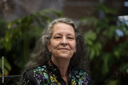 A smiling person with long silver gray hair and face piercings expresses their self-described queer gender identity and being middle aged. They are in their garden, a place they love.