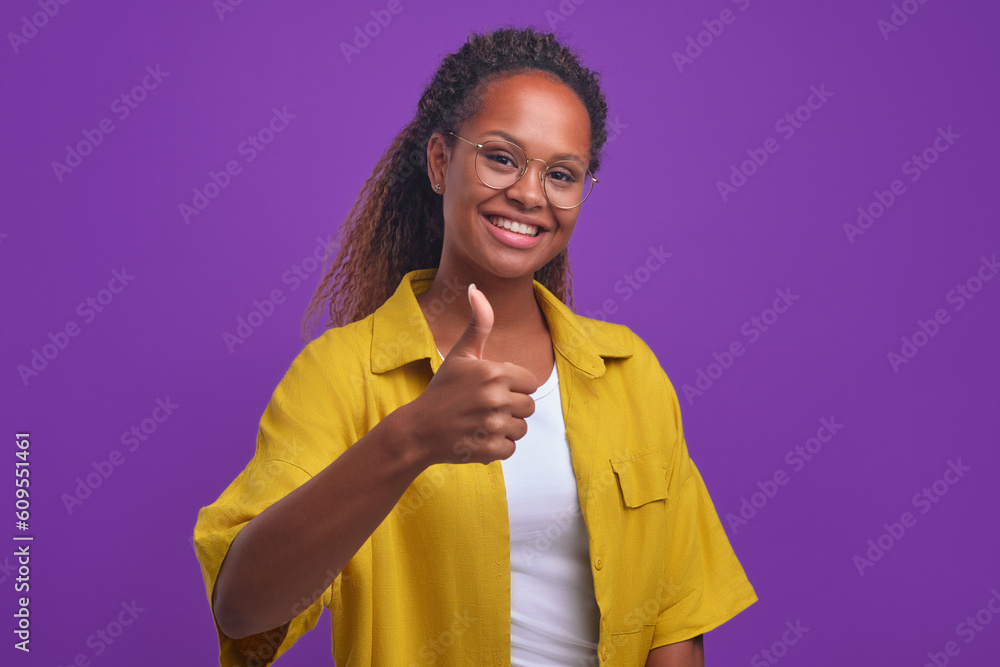 Young successful ethnic African American woman generation z in casual clothes gives thumbs up and looks at camera with smile recommending cool product at bargain price stands in purple studio. Young successful ethnic African American woman generation z in casual clothes gives thumbs up and looks at camera with smile recommending cool product at bargain price stands in purple studio.