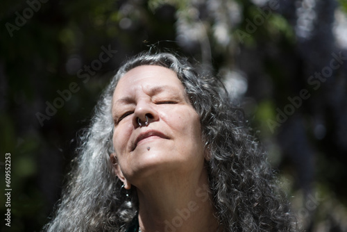 With their eyes closed, an older LGBTQ person has their head upturned, basking in the warm sunlight. Their long silver hair flows past their shoulders, and their face piercings glimmer.