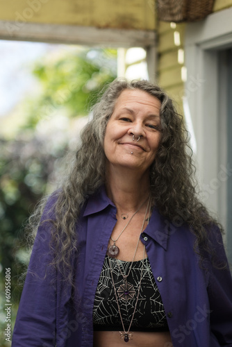 Smiling joyfully, an LGBTQ person poses in their backyard. They have a purple top, chunky jewelry, and nose and chin piercings. Their long silver hair is a testament to their maturity.