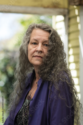 Looking over their shoulder, an older LGBTQ person looks seriously at the camera. They have long silver hair, face piercings, and chunky jewelry. They are at peace in their backyard.