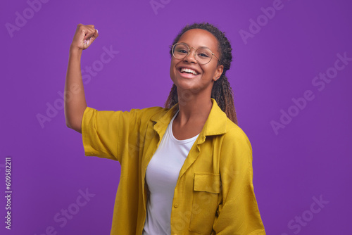 Tableau sur toile Young funny African American woman in casual clothes showing biceps on arm and laughing showing off modest achievements after going to fitness club or calling for sports stands in purple studio