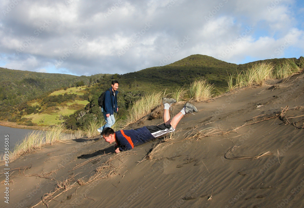 Teenage boy sliding down sand dunes with father looking on. Father and son playing together. Lake Wainamu sand dunes at Bethells beach. Auckland.