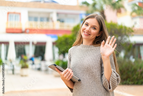 Young pretty blonde woman holding a tablet at outdoors saluting with hand with happy expression