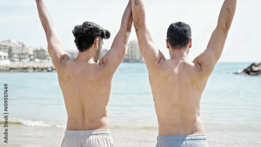 Two men tourist couple standing backwards with hands raised up at beach Two men tourist couple standing backwards with hands raised up at beach