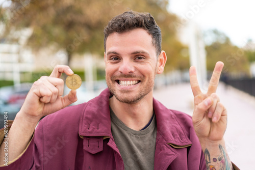 Young caucasian man holding a Bitcoin at outdoors smiling and showing victory sign