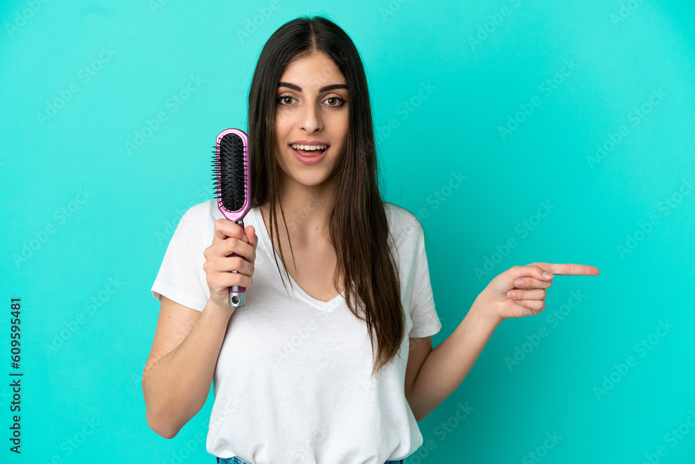 Young caucasian woman with hair comb isolated on blue background surprised and pointing finger to the side Young caucasian woman with hair comb isolated on blue background surprised and pointing finger to the side