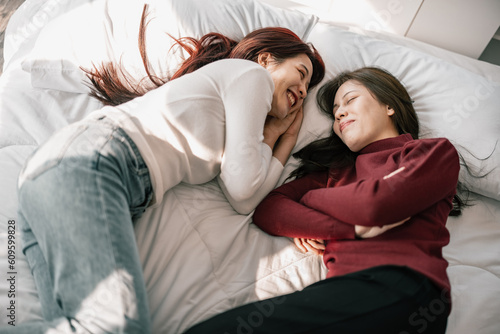 Asian lesbian partners smile, relax, laugh, affectionately pinch and kiss each other's cheeks while lying on the bed. A combination of personal charm, a good sense of humor, and a romantic moment.