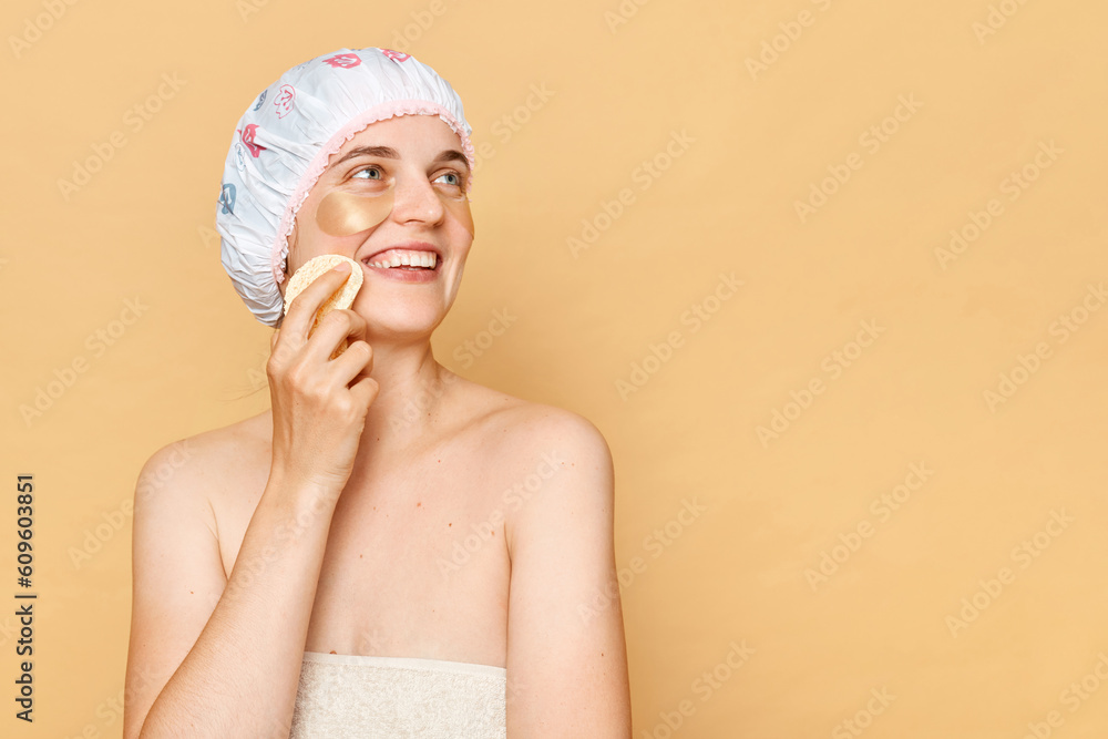 Cheerful young female model has pleasant smile,holding cosmetic sponge, enjoys all benefits of patches, reduces wrinkles, wears wrapped towel and shower cap on head. Cheerful young female model has pleasant smile,holding cosmetic sponge, enjoys all benefits of patches, reduces wrinkles, wears wrapped towel and shower cap on head.