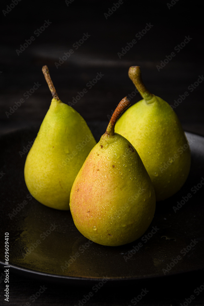 Close-up of three green pears with water drops in black plate on dark table, vertical, with copy space