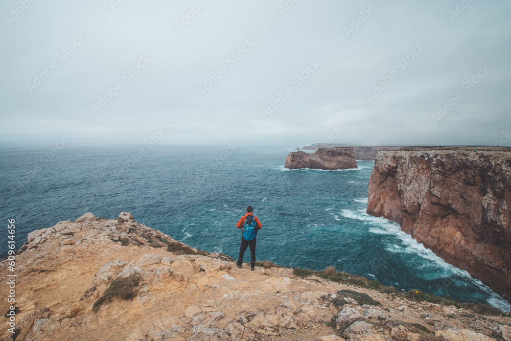 Fototapeta premium Black-haired adventurer stands at the end of Cape Cabo de Sao Vicente in the southwest of Portugal in the Algarve region. Man is enjoying his freedom. Wandering of Fisherman Trail
