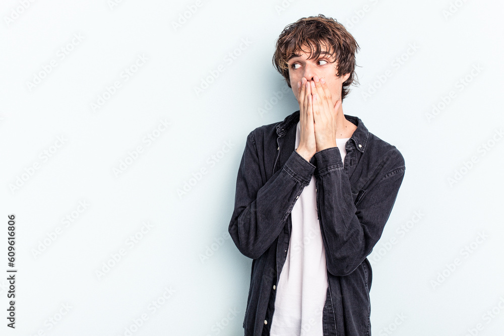 Young caucasian man isolated on blue background thoughtful looking to a copy space covering mouth with hand. Young caucasian man isolated on blue background thoughtful looking to a copy space covering mouth with hand.