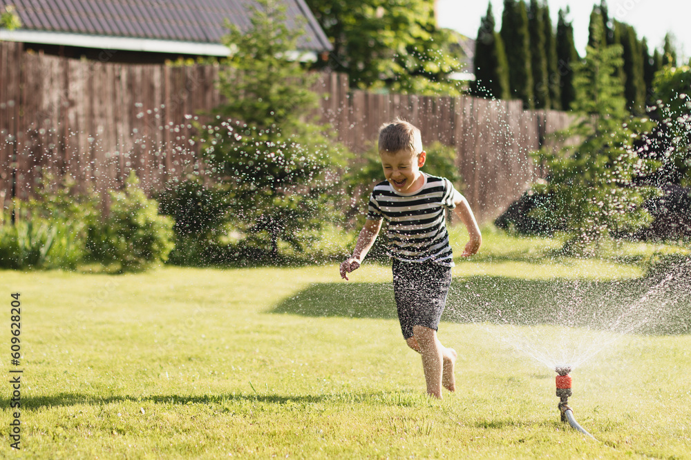 A little wet boy runs barefoot on the lawn next to the sprinkler. The concept of a happy, carefree childhood and vacation - obrazy, fototapety, plakaty 