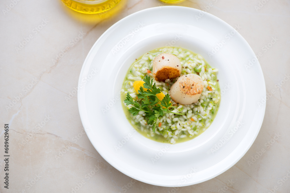 White plate with green pea risotto and seared scallops, elevated view on a beige marble background, horizontal shot White plate with green pea risotto and seared scallops, elevated view on a beige marble background, horizontal shot