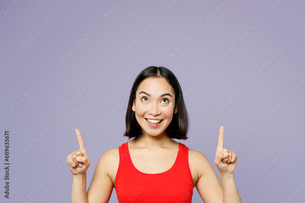 Young woman of Asian ethnicity she wear casual clothes red tank shirt point index finger overhead indicate on workspace area copy space mock up isolated on plain pastel light purple background studio