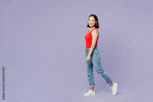 Full body sideways happy young woman of Asian ethnicity she wear casual clothes red tank shirt walk go look camera isolated on plain pastel light purple background studio portrait. Lifestyle concept.