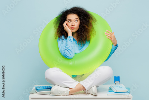 Full body young employee business woman wear casual shirt sit work at white office desk dream about free time vacations hold inflatable ring isolated on plain pastel blue background studio portrait. #609634090