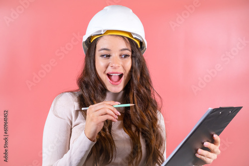 Woman wearing white hardhat writes information for building project on clipboard. Brown-haired architect with smiling and delighted expression enjoys job