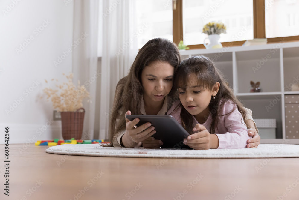 Focused young mom and serious little kid girl resting close on heating floor, using educational app on tablet, playing online learning game, reading book on screen, enjoying family leisure Focused young mom and serious little kid girl resting close on heating floor, using educational app on tablet, playing online learning game, reading book on screen, enjoying family leisure