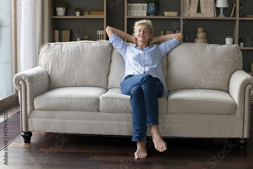 Positive sleepy mature homeowner woman stretching body on soft comfortable couch, relaxing at cozy home, enjoying comfort, relaxation, daydreaming, leisure in modern apartment. Full length shot