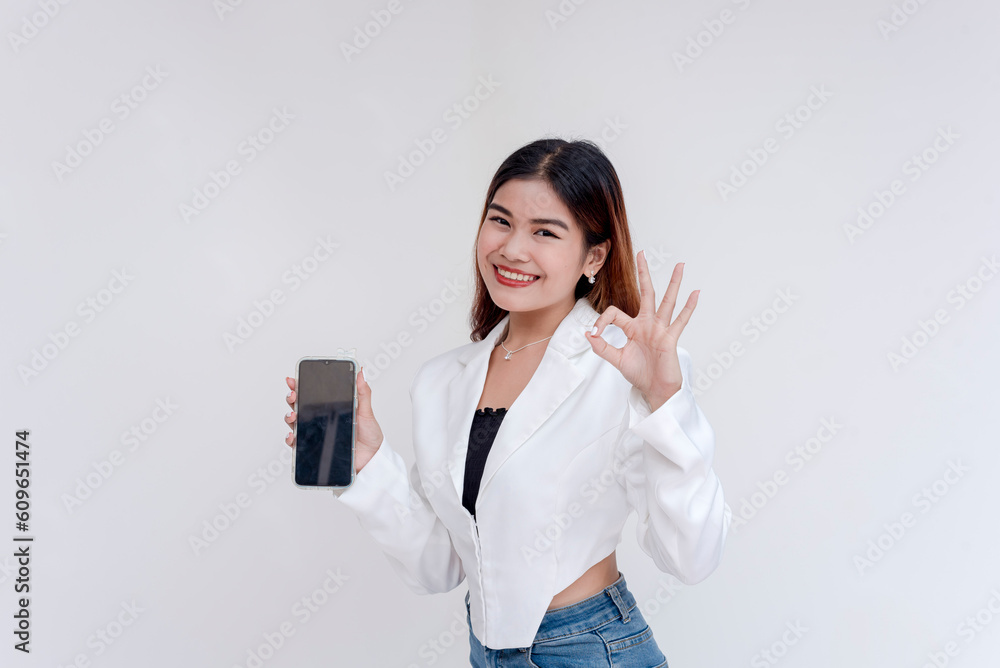 A happy and smiling young woman posing with a smart phone in her hand while doing an okay gesture. Isolated on a white background. A happy and smiling young woman posing with a smart phone in her hand while doing an okay gesture. Isolated on a white background.