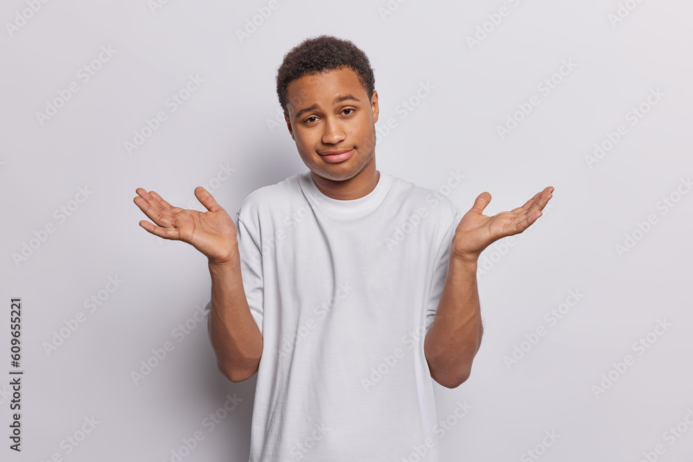 Waist up shot of dark skinned man expresses uncertainty and faces dilemma spreads palms dressed in casual t shirt isolated over white wall. Questioned African guy expresses thoughtful consideration Waist up shot of dark skinned man expresses uncertainty and faces dilemma spreads palms dressed in casual t shirt isolated over white wall. Questioned African guy expresses thoughtful consideration