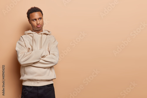 Dejected dark skinned man gazes aside with folded arms frowns with displeasure has somber expression reveals sense of hurt and offence dressed in hoodie and black jeans isolated over brown background