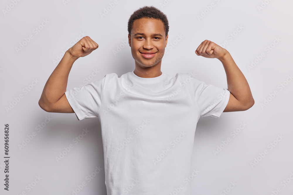 Strong African man raises arms shows biceps muscles wears casual tshirt poses against white background. Smiling guy being in great physical condition working out in gym bregging about achievements