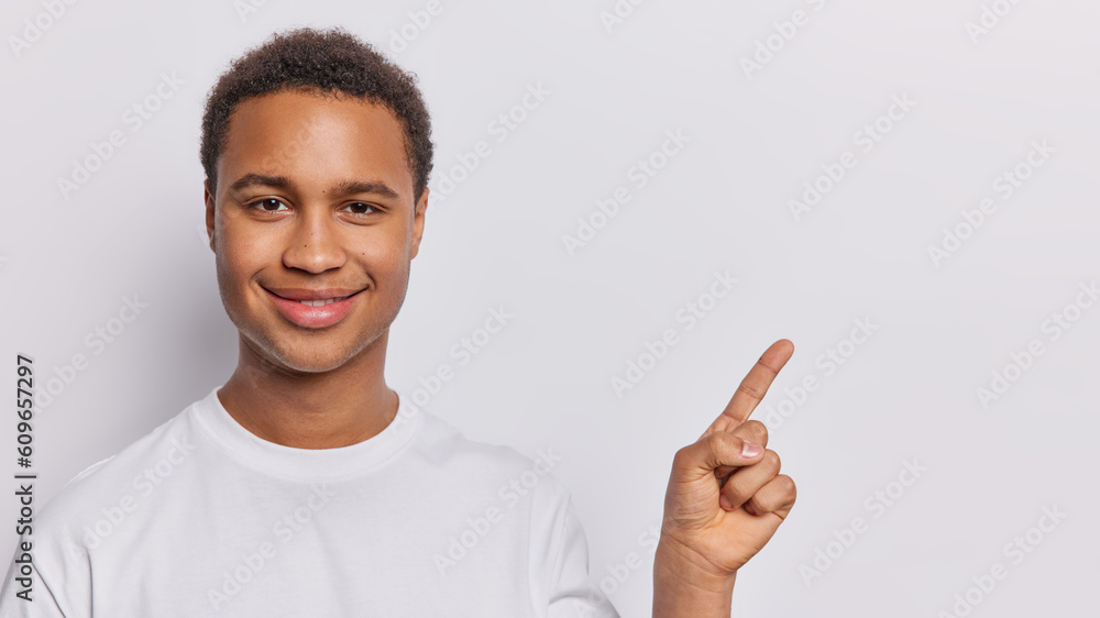 Cheerful dark skinned man promots product that demands discovery smiles pleasantly points index finger on copy space gives advice or advertises product dressed in casual t shirt isolated on white wall Cheerful dark skinned man promots product that demands discovery smiles pleasantly points index finger on copy space gives advice or advertises product dressed in casual t shirt isolated on white wall