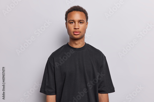 Portrait of serious dark skinned young man looks with calm self confident expression has determined look wears casual black t shirt isolated over white background listens important information