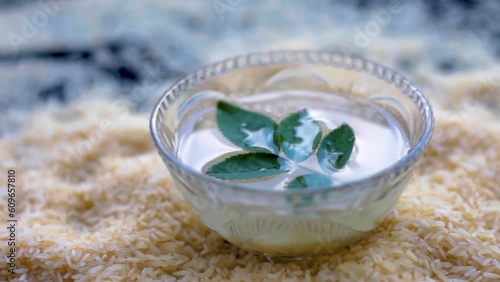 Oryza sativa and Oryza glaberrima or raw lachkari kolam rice grains on a black surface along with a glass bowl containing rice water in it with some rose leaves on top, used in beauty products and fac photo