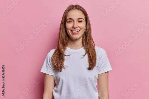 Portrait of lovely European millennial girl with straight hair smiles toothily being in good mood dressed in casual white t shirt isolated over pink studio background. Positive human emotions