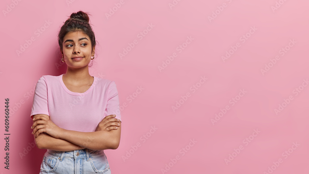 Horizontal shot of young Indian girl with hair bun keesps arms folded stands in thoughtful pose concentrated aside tries to make decision dressed in casual t shirt and jeans pink background. Horizontal shot of young Indian girl with hair bun keesps arms folded stands in thoughtful pose concentrated aside tries to make decision dressed in casual t shirt and jeans pink background.