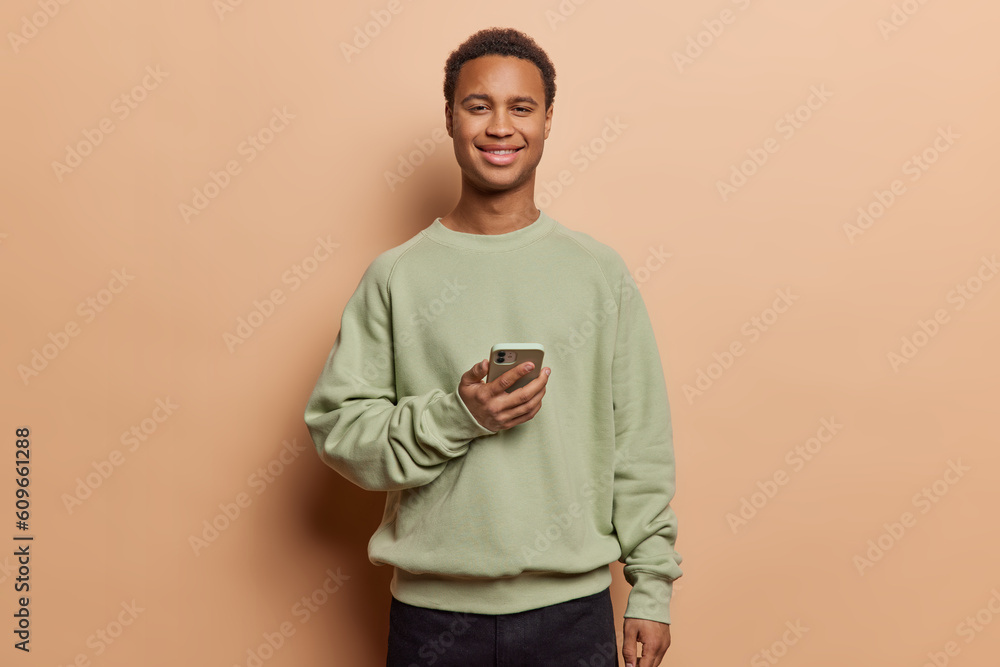 Dark skinned man wears pullover holds mobile phone his expression filled with satisfaction as he checks notification exemplifying harmonious coexistence of technology and fashion in modern lifestyle. Dark skinned man wears pullover holds mobile phone his expression filled with satisfaction as he checks notification exemplifying harmonious coexistence of technology and fashion in modern lifestyle.