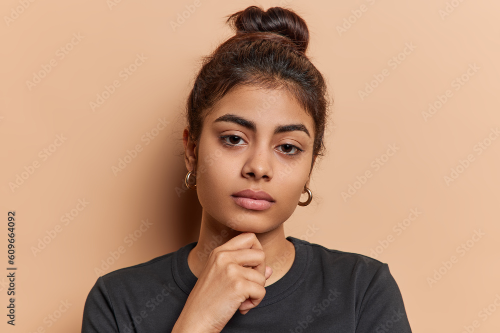 Headshot of lovely Indian woman keeps hand under chin focused directly at camera has calm expression dressed in casual black t shirt isolated over brown background. Human facial expressions. - obrazy, fototapety, plakaty 