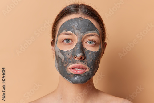 Daily skincare routine concept. Young dark haired European woman applies clay face mask indulging in moment of self care and rejuvenation isolated over brown background pampers skin poses indoor