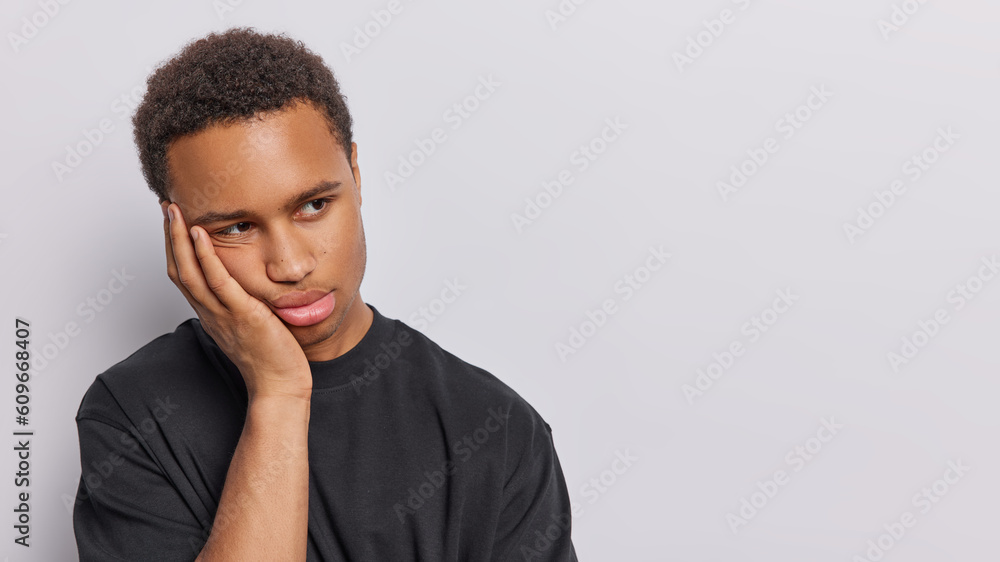 Horizontal shot of frustrated bored dark skinned man keeps hand on cheek looks discontent aside has nothing to do dressed in casual black t shirt isolated over white background copy space for promo Horizontal shot of frustrated bored dark skinned man keeps hand on cheek looks discontent aside has nothing to do dressed in casual black t shirt isolated over white background copy space for promo