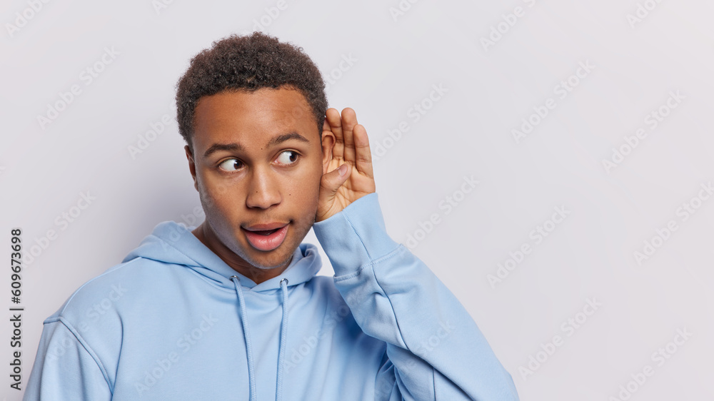 Surprised dark skinned man tries to overhear someone holds hand near ear tries to understand words eavesdropping has stunned expression wears blue sweatshirt isolated over white background copy space Surprised dark skinned man tries to overhear someone holds hand near ear tries to understand words eavesdropping has stunned expression wears blue sweatshirt isolated over white background copy space
