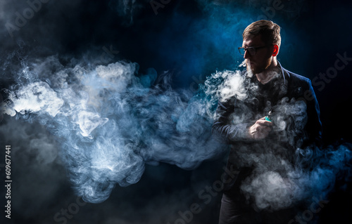Man in smoke from vaping. Unrecognizable vaper guy. Human vaping while standing in dark. Vaper blows steam out nose. Man with electronic gadget for vaping. E-cig in hand male. Electronic cigarettes © Grispb