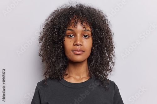 Portrait of serious curly haired teenage girl looks directly at camera has calm relaxed expression wears casual black t shirt isolated over white wall. Attractive female model poses for making photo