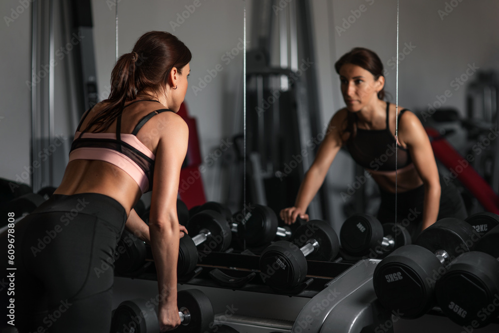 Young woman takes dumbbells in the gym and looks in the mirror Young woman takes dumbbells in the gym and looks in the mirror