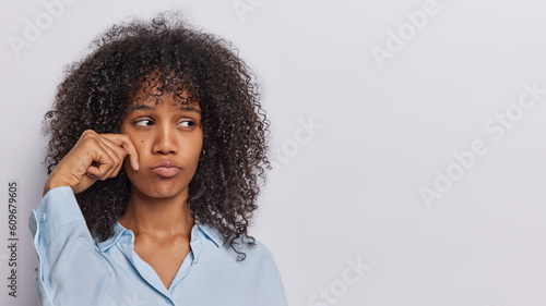 Upset curly haired African woman with displease sulking expression wipes tears has dissatisfied expression wears formal shirt isolated over white background copy space for your advertisement