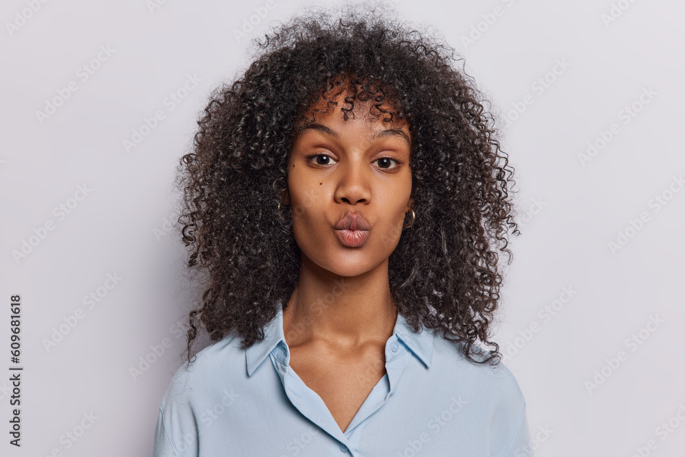 Portrait of good looking young woman with curly hair keeps lips rounded looks with romantic expression focused at camera wears formal shirt isolated over white background. Human face expressions Portrait of good looking young woman with curly hair keeps lips rounded looks with romantic expression focused at camera wears formal shirt isolated over white background. Human face expressions