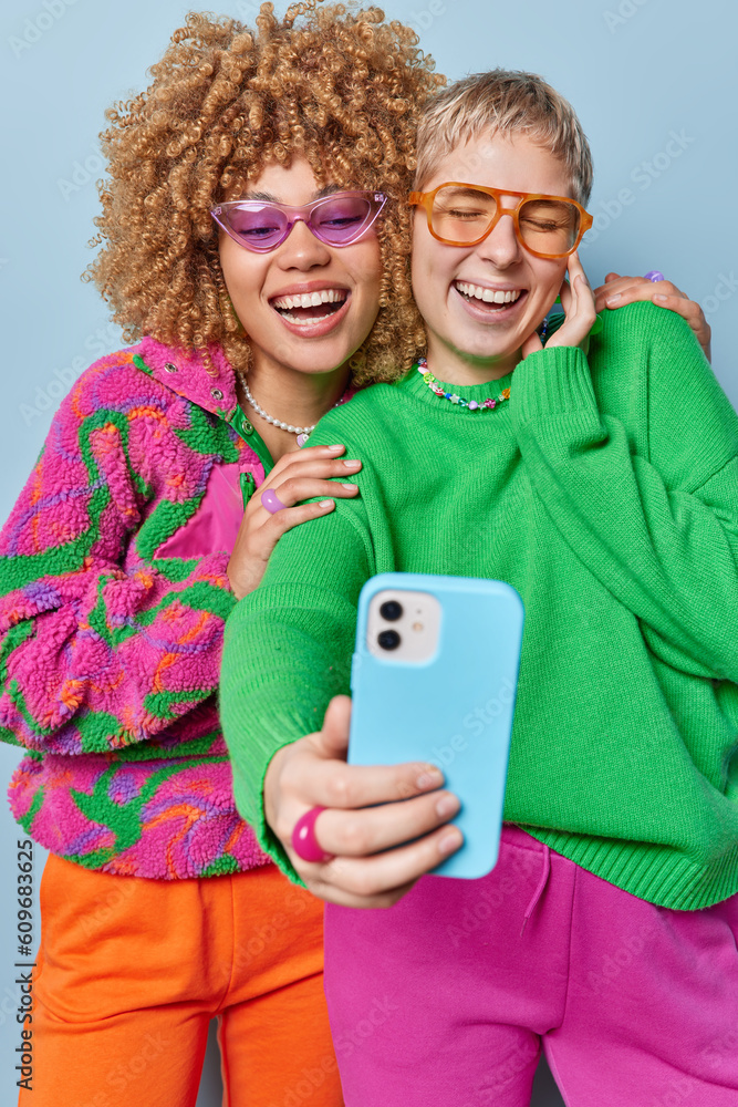 © wayhome.studio  - Vertical shot of overjoyed stylish women have fun make selfie via smartphone dressed in casual colorful clothes laughs at something funny stand next to each other isollated on blue studio wall