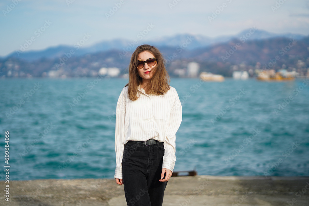 Beautiful girl walking near sea. Young woman with casual clothes standing near water. Seascape with mountains. Walk on concrete embankment. Girl wearing sunglasses smiles and looks straight at camera
