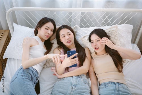 Top view of beautiful young Asian women lying on bed watching smartphone together and talking at home. Group of korean cheerful female friends having fun together on the weekend. Friendship concept © edward_indy