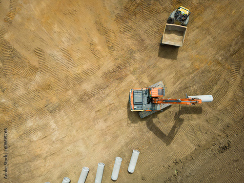 UAV view of an excavator seen taking part of a water pipe from a dumper truck. Seen ready to be dug into the ground, ready for a future English housing estate.