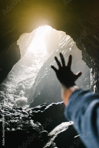 A man holds a silouetted hand out toward the entrance of a cave with water and sunlight streaming in photo